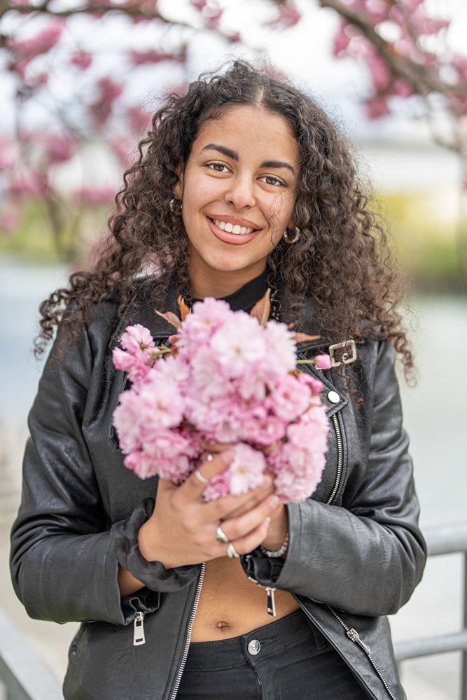 Portrait einer jungen Frau mit Locken und Lederjacke, die einen Strauß rosafarbener Kirschblüten hält – natürliches Outdoor-Modefoto von Der mobile Fotograf & Videograf in Villach, Kärnten