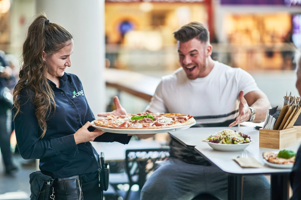 Servicekraft serviert eine große Pizza an einen lachenden Gast im Restaurant Al Dente im Atrio Villach; daneben steht ein Salat.