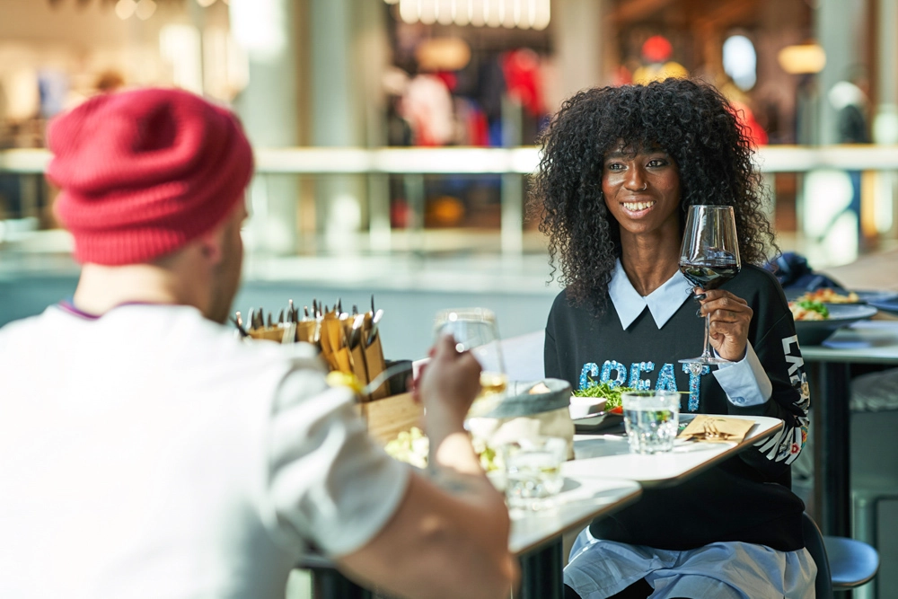 Gästin hebt ein Glas Rotwein beim Essen im Restaurant Al Dente im Atrio Villach; Mall-Atmosphäre im Hintergrund.