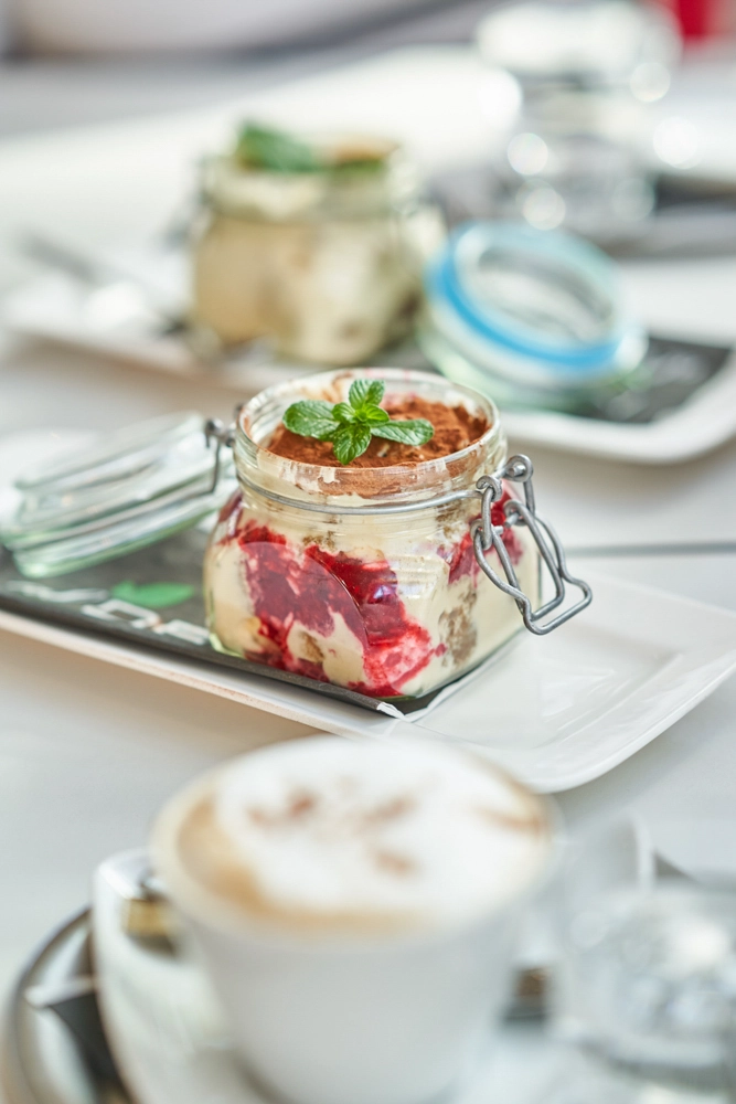 Dessert im Weckglas mit Beeren und Minze auf Teller; unscharf im Vordergrund eine Cappuccinotasse.