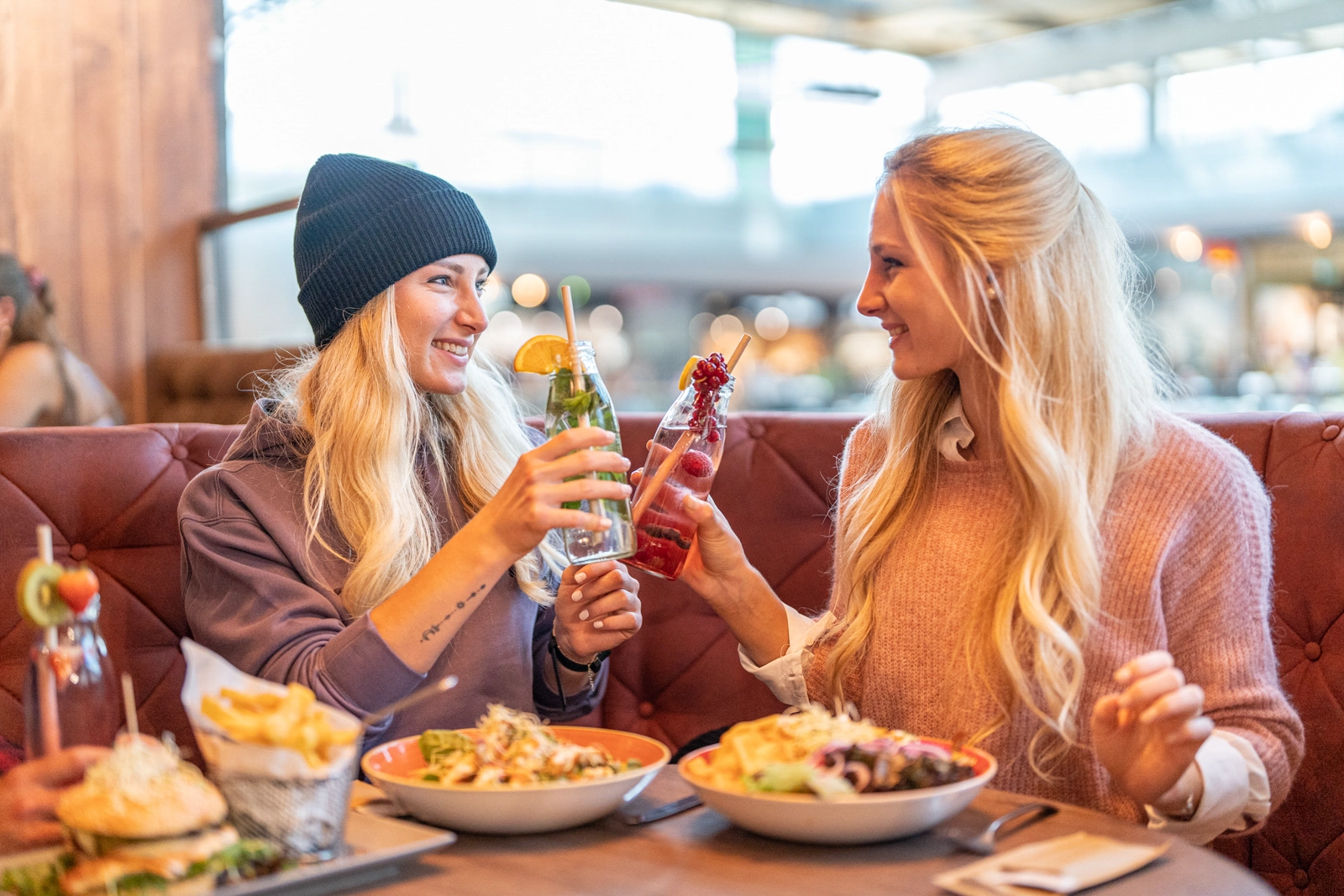 Foodfotografie – Alexandra und Viktoria Sedlmaier stoßen mit Limonadenflaschen an; vor ihnen Salate, Burger, Bowls und Pommes im Franz Streetfood Restaurant im Atrio Villach.
